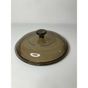 Puede incluir: Una tapa de vidrio marrón con un pomo redondeado. La tapa es circular y tiene un borde ligeramente elevado. El vidrio es transparente, permitiendo el paso de la luz. El pomo es de un tono marrón más oscuro.