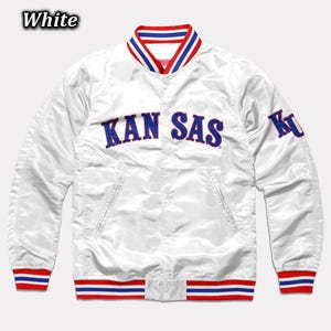 May include: White satin jacket with the word "KANSAS" in blue block letters across the chest. The jacket has red and blue striped trim on the collar, cuffs, and waist. The letter "K" is on the left sleeve. The word "White" is in the top left corner.
