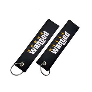 May include: Two black rectangular fabric keychains featuring the word wanted embroidered in white capital letters. Each keychain is decorated with four small yellow stars arranged in a vertical line above the text and includes a silver metal ring.