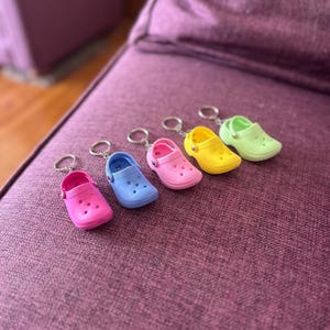 May include: A collection of miniature, colorful clogs keychains in pink, blue, pink, yellow, and green. Each keychain features a small, detailed clog with a silver-colored keyring attached. The clogs are arranged in a row on a purple surface.