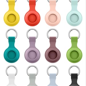 Op de afbeelding: Een verzameling van twaalf kleurrijke AirTag-houders in verschillende tinten, waaronder geel, rood, roze en groen. Elke houder heeft een cirkelvormig ontwerp met een sleutelringbevestiging. De houders zijn gemaakt van een zacht, flexibel materiaal.