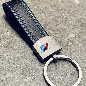 May include: Black leather keychain with blue, red, and white stitching. The keychain features a silver-colored metal clasp and a metal emblem with the letters "M" and the BMW logo in blue, red, and white.