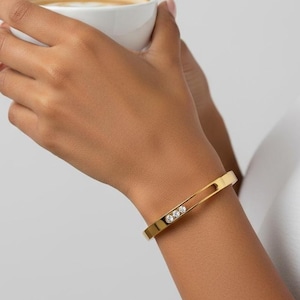 May include: A gold-coloured bangle bracelet featuring three clear, round gemstones. The bracelet has a rectangular shape with a cut-out design. The person is holding a white cup of coffee.