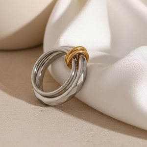May include: A silver-colored ring with a geometric design, featuring a gold-colored band intertwined. The ring is displayed on a white fabric, with a neutral background. This piece of jewelry is suitable for everyday wear.