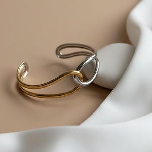 May include: A two-tone cuff bracelet. The bracelet features a gold-colored band that curves around a silver-colored oval. The bracelet is displayed on a white satin cloth against a neutral background.