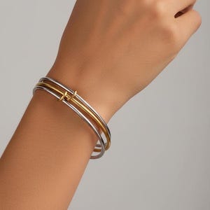 Peut inclure: Ensemble de trois bracelets joncs argentés et dorés superposés sur un poignet. Le bracelet supérieur présente un détail de nœud doré. Fond de couleur neutre.