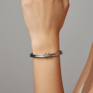 May include: A silver and gold-tone bracelet. The bracelet features a thick, rounded design with two parallel silver bands and two small gold-tone rings. The jewelry is worn on a wrist against a neutral gray background.