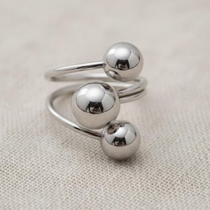 May include: A silver-colored ring with a spiral design. The ring features three spherical, reflective accents. The ring is made of a shiny, metallic material and sits on a neutral-toned fabric surface. The ring is a piece of jewelry.