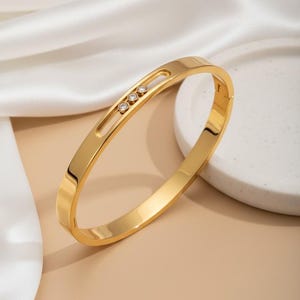 May include: A gold-coloured bangle bracelet featuring three small, clear gemstones set within a rectangular cutout. The bracelet has a smooth, polished finish and a simple, elegant design. The background is a soft beige colour.