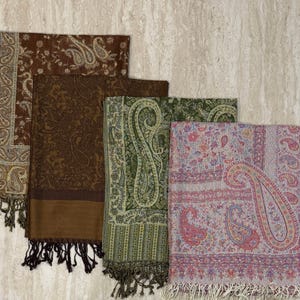 May include: An assortment of paisley-patterned scarves in shades of brown, green, and pink. Each scarf features a detailed design and fringe along the edges. The scarves are folded and stacked, showcasing their intricate patterns and textures.