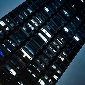 Könnte beinhalten: Niedrigwinkelaufnahme eines hohen Wolkenkratzers bei Nacht. Die Fassade des Gebäudes ist ein Raster aus dunklen Fenstern, von denen viele durch Innenbeleuchtung beleuchtet werden. Der Himmel ist tiefblau und bildet einen Kontrast zur dunklen Außenfassade des Gebäudes.