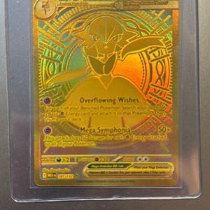 Könnte beinhalten: Eine goldfarbene Mega Gardevoir EX Sammelkarte in einer Schutzhülle. Die Karte zeigt eine stilisierte Illustration von Gardevoir mit Text wie "Overflowing Wishes" und "Mega Symphonia". Ein Sammlerstück.