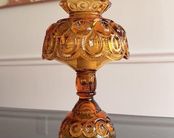 Puede incluir: Una lámpara de cristal color ámbar con un patrón prominente de lunas y estrellas en la pantalla y la base. La lámpara tiene un borde festoneado, un tallo central tipo pedestal y una base circular ancha, hecha de vidrio prensado translúcido.