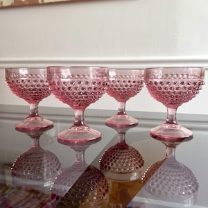 May include: Four pink hobnail wine glasses with a textured surface. The glasses have a round base and a stem. The glasses are reflected on a glass surface.
