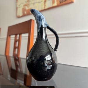 May include: A black ceramic pitcher with a curved spout and handle. The top of the pitcher is a dark blue color. The pitcher has a glossy finish and is sitting on a reflective surface. The pitcher is a decorative item.
