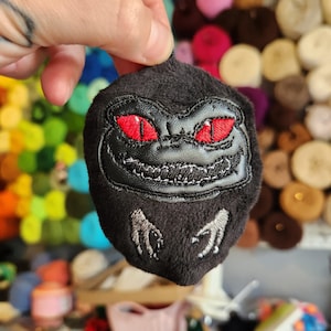May include: A black plush Critters-themed keychain with red eyes and a stitched leather face. The keychain has embroidered details and small, clawed hands. The background is blurred, showing colorful yarn and other craft supplies.