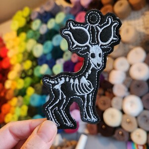以下が含まれることがあります： 黒と白の刺繍パッチ。鹿の骨格の形をしており、鹿の骨格構造、角、顔の特徴が詳細に描かれています。衣服やアクセサリーに縫い付けることを目的としたパッチです。
