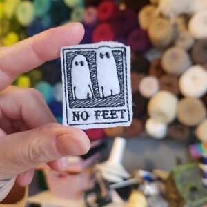 Peut inclure: Un petit patch rectangulaire en feutre blanc avec un motif brodé noir. Le motif représente deux fantômes de dessin animé et le texte "NO FEET" en gras, sans empattement. Le patch est tenu par une main.