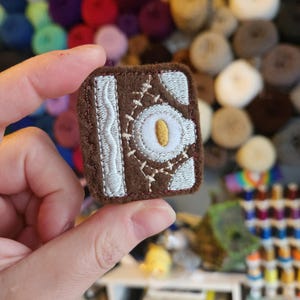 May include: A brown, square-shaped embroidered patch resembling a spellbook. The patch features white stitching around the edges and a central eye design with a yellow iris. A white, wavy line is on the left side. The patch is held in a person's hand.
