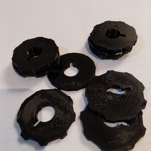 Può includere: Una collezione di oggetti circolari neri con un foro centrale e un bordo esterno dentellato. Gli oggetti sono impilati e sparsi, con testo visibile su alcuni. Gli articoli sembrano essere fatti di plastica dura.