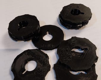 May include: A collection of black, circular objects with a central hole and a notched outer edge. The objects are stacked and scattered, with text visible on some of them. The items appear to be made of a hard plastic material.