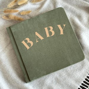 May include: A sage green baby book with the word "BABY" in a light peach color. The book is lying on a light gray blanket with a textured pattern. Dried wheat stalks are in the upper left corner.