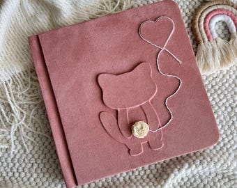 May include: A square, dusty rose-colored album with a soft, suede-like texture. The cover features an embossed outline of a cat holding a white, fabric flower. A thin, white string extends from the flower, forming a heart shape above the cat.
