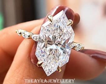 Op de afbeelding: Een gouden ring met een grote, langwerpige, zeshoekig geslepen middensteen met een briljant gefacetteerd oppervlak. De band is aan weerszijden versierd met kleine, ronde, heldere stenen. De tekst THEAYRAJEWELLERY is zichtbaar op de ringzetting.