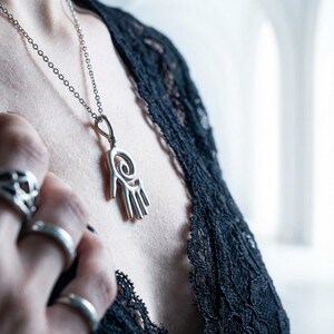 May include: Silver pendant necklace featuring a stylized hand design with a spiral and vertical lines. The pendant hangs on a delicate chain. The image also shows several silver rings and a black lace top.