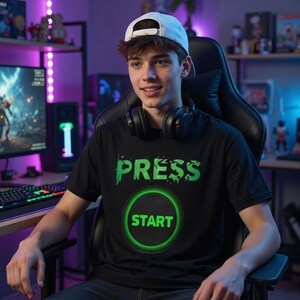 Op de afbeelding: Zwart T-shirt met de groene tekst "PRESS START". Het shirt heeft een groene cirkel met daarin het woord "START". De persoon op de foto draagt een witte baseball cap en een zwarte koptelefoon.