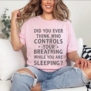 May include: A light pink t-shirt with the text "DID YOU EVER THINK WHO CONTROLS YOUR BREATHING WHILE YOU ARE SLEEPING?" in gray. The shirt is worn by a person wearing ripped blue jeans.