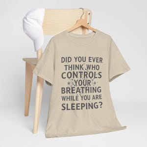 Puede incluir: Una camiseta beige con el texto "DID YOU EVER THINK WHO CONTROLS YOUR BREATHING WHILE YOU ARE SLEEPING?" en gris oscuro. Una gorra de béisbol blanca descansa sobre una silla de madera.