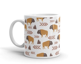 May include: White ceramic mug with a repeating pattern of brown and orange bison, brown mountains, and red arrows.