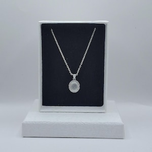 May include: A silver necklace with a circular pendant, presented in a white box with a black interior. The pendant is encircled by small, sparkling stones. The necklace is on a white textured surface.