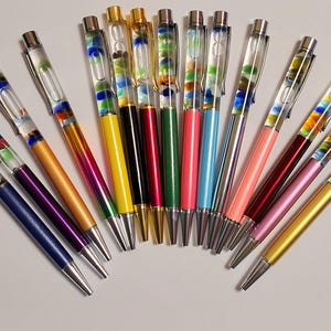 Op de afbeelding: Een verzameling kleurrijke pennen met doorzichtige vaten gevuld met decoratieve elementen. De pennen hebben zilveren accenten en zijn verkrijgbaar in verschillende kleuren, waaronder blauw, paars, goud, roze, groen en geel. De pennen zijn in een halve cirkel gerangschikt.