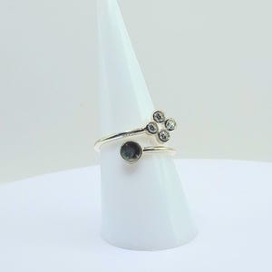 Puede incluir: Un anillo plateado con un diseño único. El anillo presenta un grupo de cuatro piedras claras y una sola piedra de color oscuro. El anillo se exhibe sobre un soporte blanco en forma de cono, resaltando su estilo elegante y moderno.