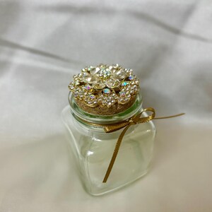 Puede incluir: Un pequeño tarro de cristal transparente con un tapón de corcho. El tapón está adornado con un diseño floral de metal dorado incrustado con pedrería transparente e iridiscente. Una cinta dorada está atada alrededor del cuello del tarro.