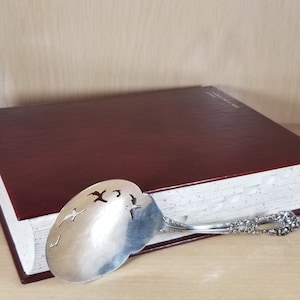 May include: A large, burgundy leather-bound book with white pages and a silver serving spoon with a decorative handle resting on top.