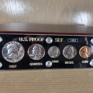 May include: A 1953 U.S. Proof Set displayed in a black case. The set includes a half dollar, quarter, dime, nickel, and cent. The coins are silver and copper-toned, with the text "U.S. Proof Set" and the year "1953" visible.