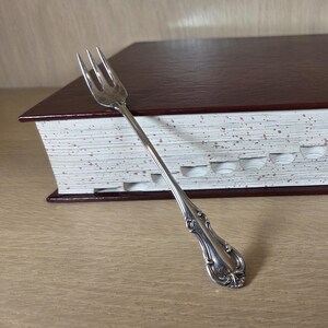 May include: A silver fork rests against a large, closed book with a dark red cover and white, speckled pages. The book has a series of stepped cutouts along its edge. The fork has an ornate handle.