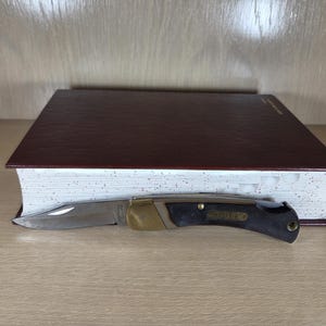 May include: A closed pocket knife with a silver blade, brass bolster, and black handle rests on a closed, burgundy-colored book. The knife is positioned horizontally, with the blade facing left.