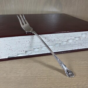 May include: A silver fork with an ornate handle rests on a closed, burgundy-colored book with white-edged pages. The fork's tines are visible, and the book has a textured cover. The background is a light-colored wooden surface.