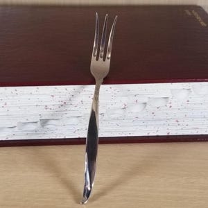 May include: A stainless steel fork with three tines, featuring a curved handle. The fork is positioned vertically against a background of a dark red book with white-edged pages. The fork is a kitchen utensil.