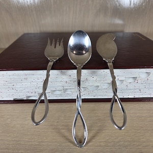May include: Three-piece silver serving set, including a fork, spoon, and spreader, with twisted handles. The utensils are arranged in front of a closed, maroon-colored book with white-edged pages. The set is likely for serving appetizers or desserts.
