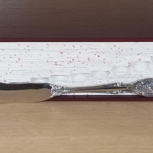 May include: A closed, hardcover book with a dark brown cover and speckled white pages. A silver butter knife is inserted between the pages, with the blade facing left. The book is resting on a light brown surface.
