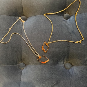 May include: Two gold-colored necklaces with pendant outlines of a state. The necklaces are on a dark gray, tufted surface. The chains are delicate and the pendants are detailed.