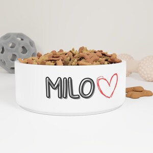 Puede incluir: Cuenco de cerámica blanca para mascotas lleno de comida para perros. El cuenco tiene el nombre "MILO" en letras negras y grises con un símbolo de corazón rojo. Otros juguetes para mascotas están en el fondo.