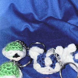 May include: Crocheted keychains in the shape of cartoon characters. The keychains are white with green and blue accents. Each keychain has a silver-colored metal ring attached.