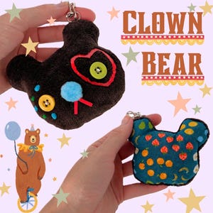 May include: A plush bear-shaped keychain with a dark brown textured front featuring a light blue pom-pom nose, yellow and green button eyes, and red stitching. The reverse side is a teal fabric patterned with strawberries, oranges, and bananas.