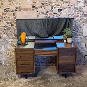 May include: A dark wood vanity desk featuring a central mirror, two side columns with three drawers each, and a central open storage space. The desk has a smooth finish with horizontal grain patterns and is supported by four short, dark, tapered legs.